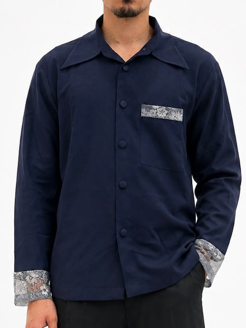 A man wearing a navy blue long-sleeve button-up shirt with contrasting gray and white patterned pocket and cuff details, paired with black pants against a plain white Close-up front view of navy oversized overshirt highlighting the floral lace chest pocket, concealed button placket, and lace cuffs.background.